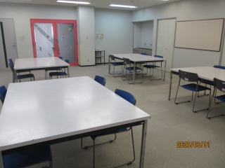 布掲示板に向かって左手にドア、左奥に流し台、中央に4人掛けの大きな机が4つ配置されている工作室を写した写真