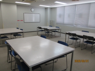 ホワイトボードに向かって右側に大きな窓、中央に4人掛けの大きなテーブルが4つ配置されている工作室を写した写真