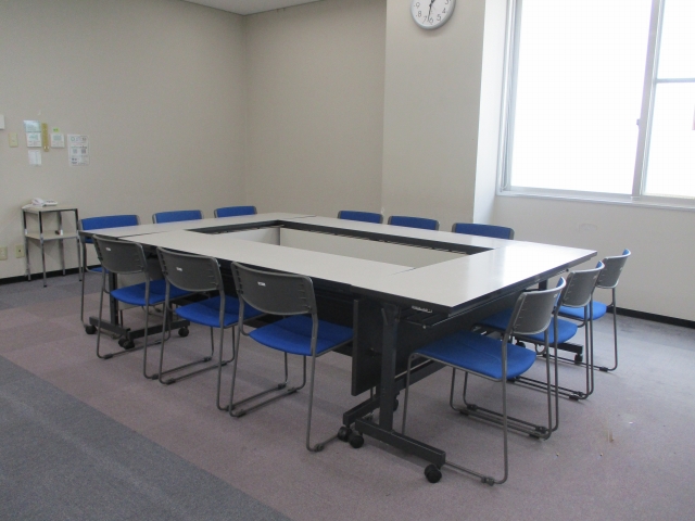 右に大きな窓があり、中央に長机を四角く囲みその周りに椅子を12脚並べた会議室 半室を写した写真