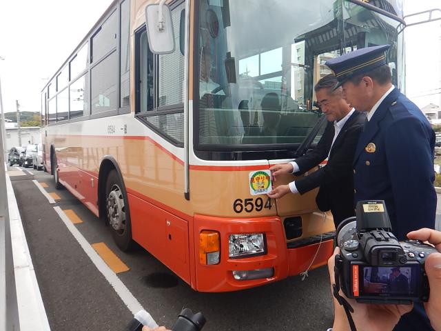 ふたりの男性がオレンジ色の大型バスの前方にステッカーを貼っている写真