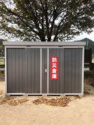大きく赤い長方形の枠に白字で「防災倉庫」と書かれた扉の物置が屋外に設置されている写真