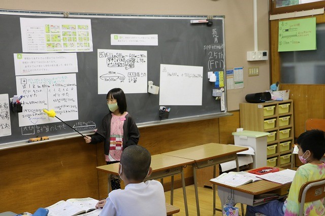 女子生徒が、黒板に貼られた白い紙の資料を差し棒でさしながら説明している様子の写真