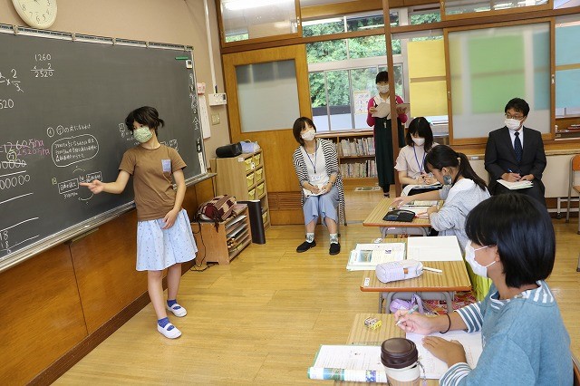 一人の女子生徒が、右手で黒板を指差してみんなの前で説明している様子の写真