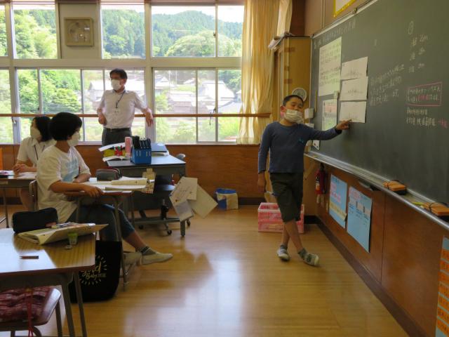 一人の男子生徒が黒板の前に立ち、黒板を指差しながら説明している様子の写真