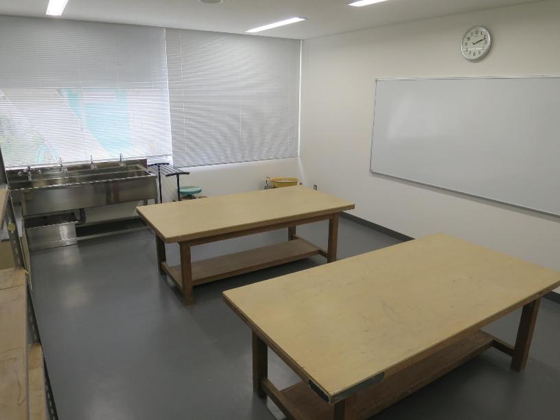 正面に流し台、右手にホワイトボード、中央に作業机が2台設けられた入り口から写した創作室の写真