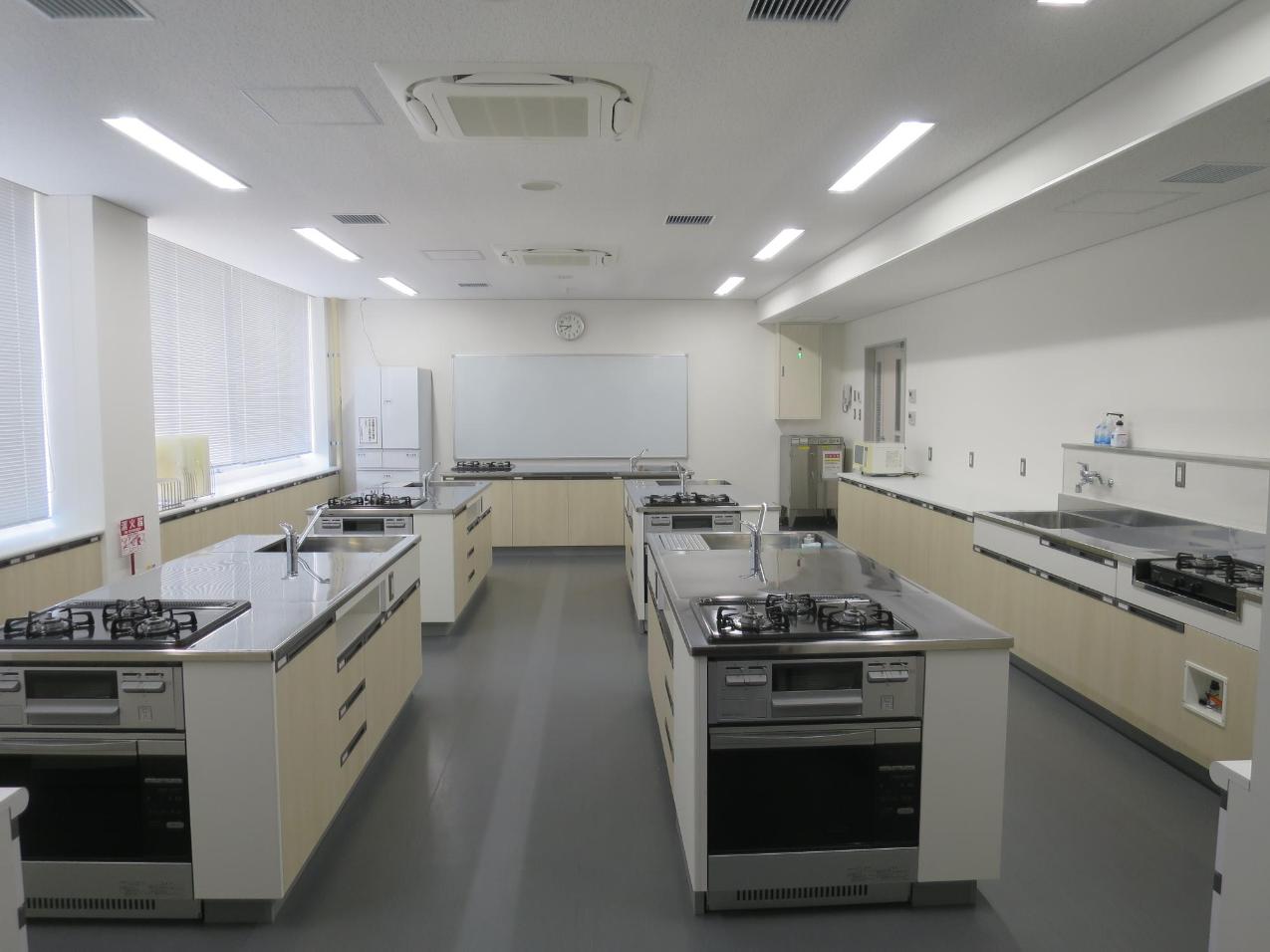 ホワイトボードに向かって右手に入り口、左手に窓、2列に並んだ調理机を4台を写した調理室の写真