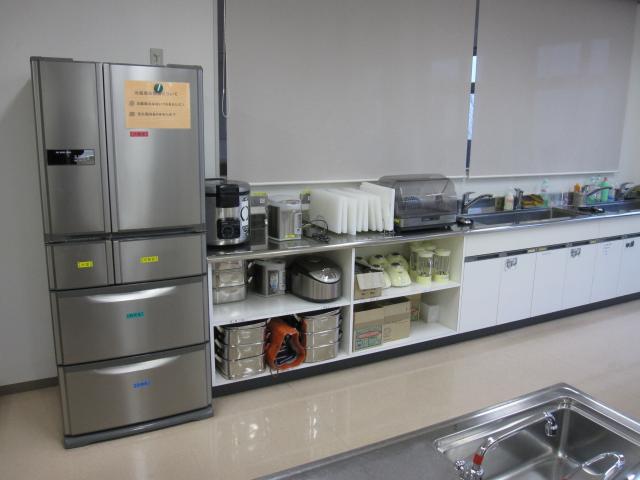 左側に冷蔵庫、中央に炊飯器などが置かれた棚、右側に流し台が写った調理室の写真