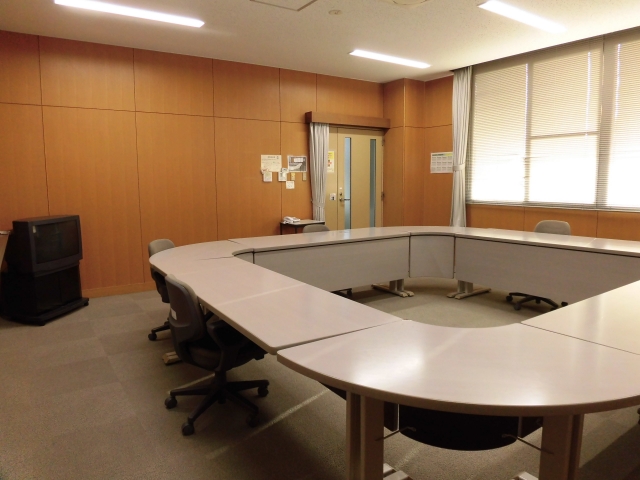 左手奥にブラウン管モニター、中央に会議室用テーブルを丸く囲みその周りに椅子を並べた様子を写した小会議室の写真