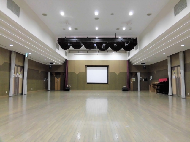 中央奥にスクリーン、左右奥に設備・備品が置かれた多目的ホールを後方から写した写真