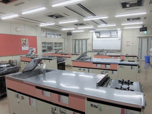 奥にホワイトボード、中央に調理台が並べられた調理室を後方から写した写真