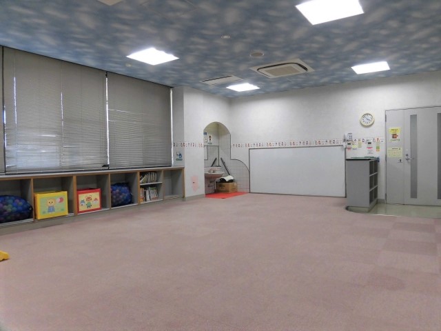 右手にドア、中央にホワイトボード、左手に窓、その下に低い棚がある幼児室を写した写真
