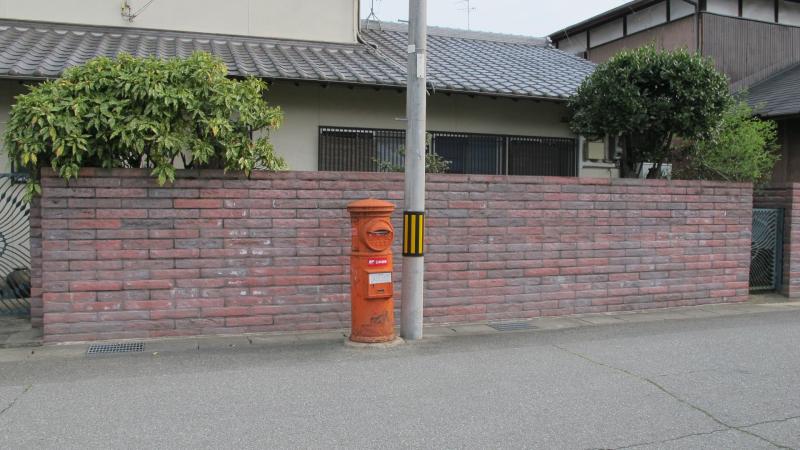 赤茶色のブロック塀の近くに設置されている丸型ポストの写真