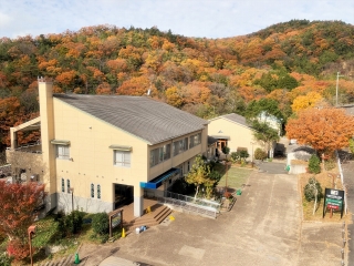 オレンジ色に紅葉した山々を背に、斜めの屋根がある大きな建物が建っている写真
