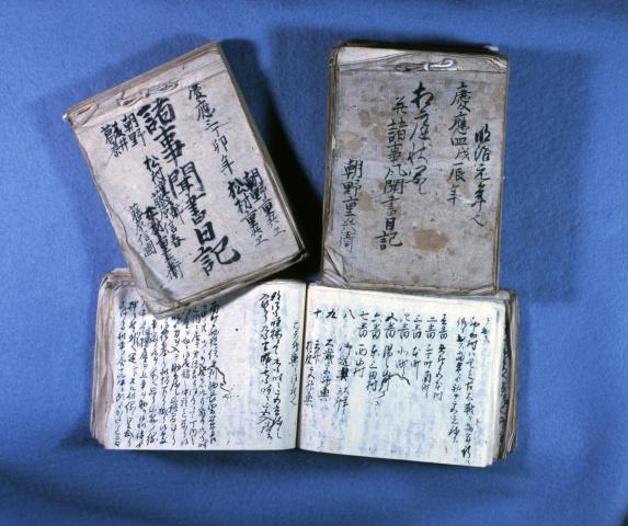 「諸事風聞記」と書かれた冊子と、年号などが書かれている日記の冊子、ページが開かれて置かれている冊子が並んでいる写真