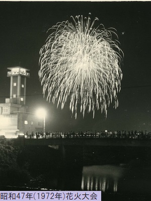 昭和47年におこなわれた花火大会で空に打ち上げられた花火を橋の上で眺める人々を遠目のアングルから撮影した白黒写真