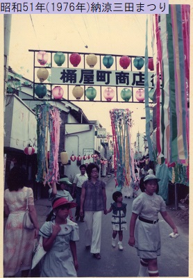 昭和51年におこなわれた納涼三田まつりで桶屋町商店街を歩く人々の頭上に掲げられた上下左右をカラフルな提灯に囲まれた商店街看板の写真
