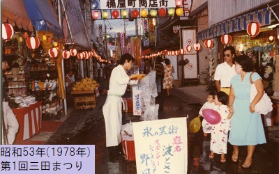 昭和53年におこなわれた第1回三田まつりで桶屋商店街の真ん中で黄色のチェーンソーを使い氷細工を作成している男性の様子を見つめる家族の写真