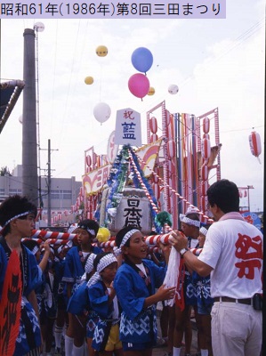 昭和61年におこなわれた第8回三田まつりでお揃いの鉢巻き法被姿でお神輿を担ぐ子どもたちと担ぎ棒に手をかけている赤文字で祭と印字されたTシャツを着た男性の写真