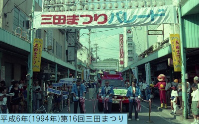 平成6年の第16回三田まつりパレードの開会式で赤文字で三田まつり水色の文字でパレードと書かれた横断幕の下でテープカットをおこなう男性関係者3人の写真