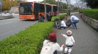 三田市内を走る連節バスオレンジアロー連を撮影しようとスタンバイしている男性スタッフ5人の後ろでバスを左手で指差す子どもの写真