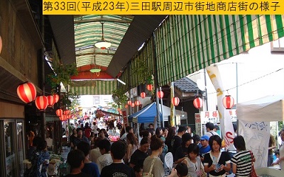 平成23年におこなわれた第33回三田まつりで人々の頭上に紅白提灯が飾られ出店が並んだ駅前商店街の様子を撮影した写真