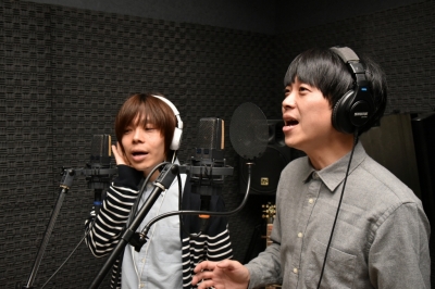 左側の男性は右手で白色のヘッドフォンに手を置き右側の男性は右手を動かしながら歌っている写真