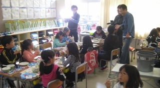 三田市立三輪小学校2年2組の教室内で机を並べて給食を食べる生徒たちの様子を撮影している男性カメラマンの左側の男性照明係と右側の映像をチェックする男性の写真