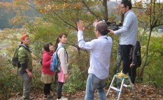 三田市にある里山高平観福の森の登山者役の女性2人男性1人に指示を出す男性とその左横で三脚の頂点に上りハンディカメラを持ちながら待機する男性カメラマンの写真