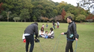 三田市内にある広々とした中央公園で遊ぶ親子連れを撮影している男性カメラマンとその右横で水色のケーブルを持った男性アシスタントの写真