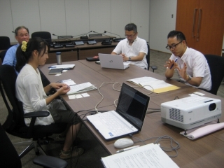 横長の会議机を2台並べ手振りを交えて三田市PRCMの意見を述べる男性とその向かい側の女性および2人の左側に座る男性2人の写真