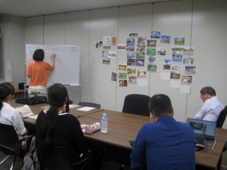 壁に貼られた三田市PRCMの資料を見ながら意見を出し合う女性2人と男性2人および資料の左側に置かれたホワイトボードに出された案を書いていく女性の写真