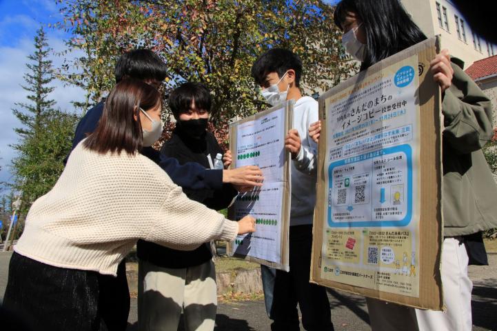 2人の子どもが持っているボードに緑色の丸いシールを貼ってシール投票している人たちの写真