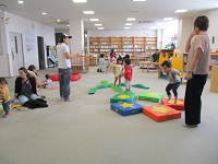 フリースペースでおもちゃの床を踏んだりしながら遊ぶ子供とそれを見守る大人の写真