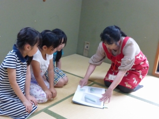 赤いエプロンの女性が3人の子どもたちに紙を見せている写真
