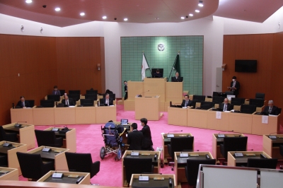 会議場の中心で席のモニターを見つめる青い車いすの人物と人物のそばにいる2人のスーツ姿の男性と会議場の奥の議席に座るスーツ姿の男性たちの写真