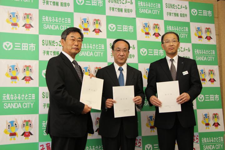 3人のメガネを掛け、黒のスーツを着た男性が締結書を前に掲げている写真