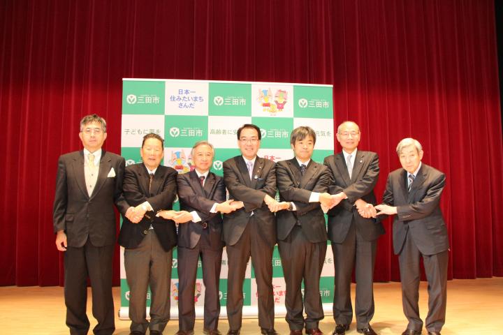 「学びの都(まち)三田」まちづくり協働宣言に臨んで、各々クロス握手を交わしつつ記念撮影に応じる森市長(写真中央)および6名の学長・校長たちの写真