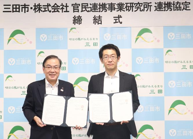 締結式にて署名した調印書を手に記念撮影に応じる、森市長と河田経営本部長の写真