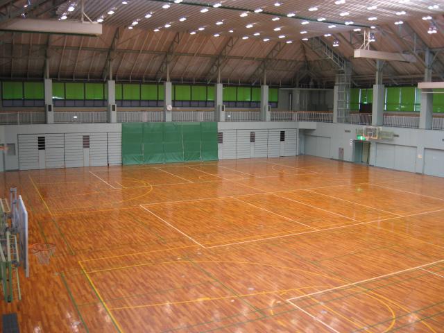 天井の多数の照明で明るくなっている、艶のある茶色い床が広がっている体育館の写真