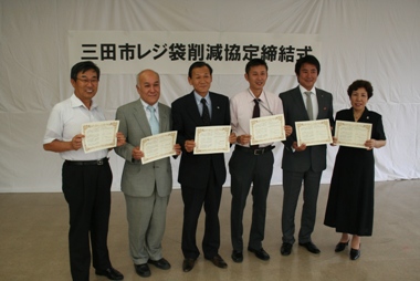 三田市レジ袋削減協定締結式と書かれた横断幕のある白い大きな布を背景に両手に賞状を持ち立つ6人の男女の写真