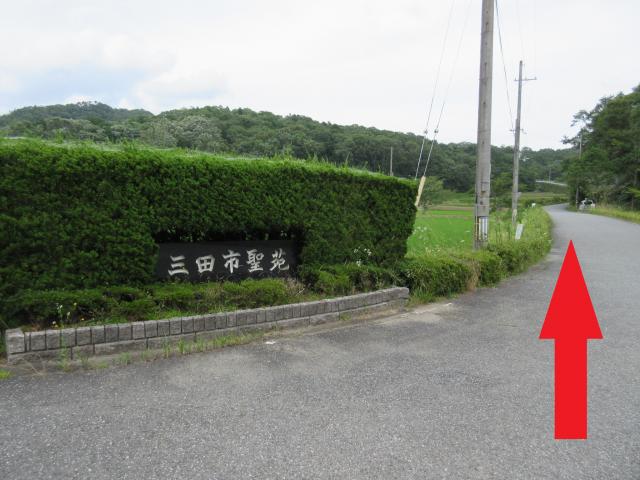 右側に道路があり、上に向いて赤い矢印が見え、左側に三田市霊苑と書かれたプレートがある写真
