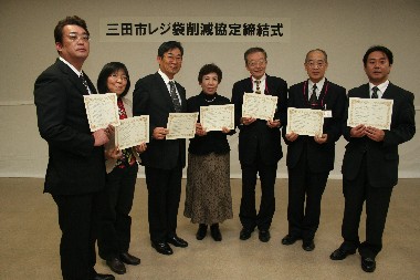 三田市レジ袋削減協定締結式と書かれた白い垂れ幕を背景に両手に賞状を持つ2人の女性と5人のスーツ姿の男性の写真