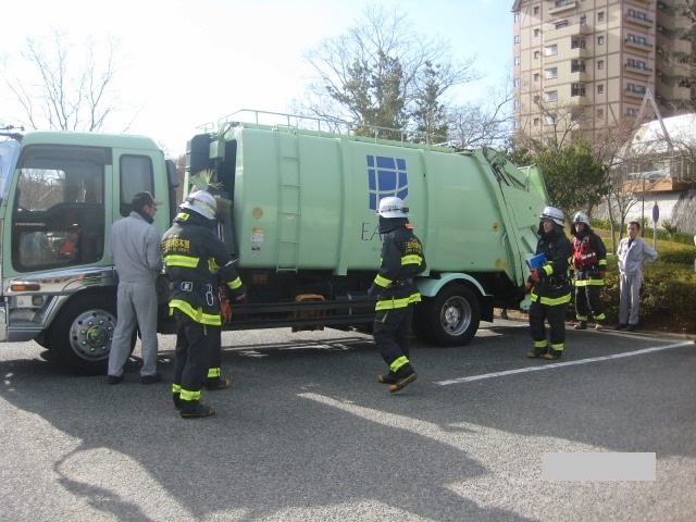 薄緑色のトラックの前に数名の作業員が立っていて消防現場検証をしている写真