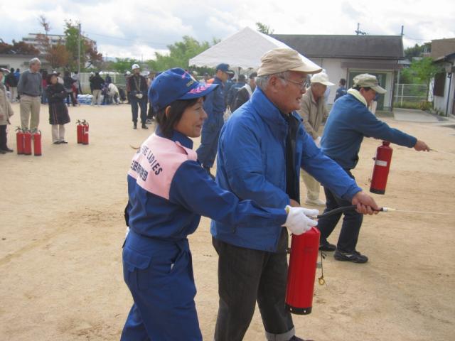 消火器を使う訓練をしている高齢の男性と、すぐ横に立って教えている女性消防団員の写真