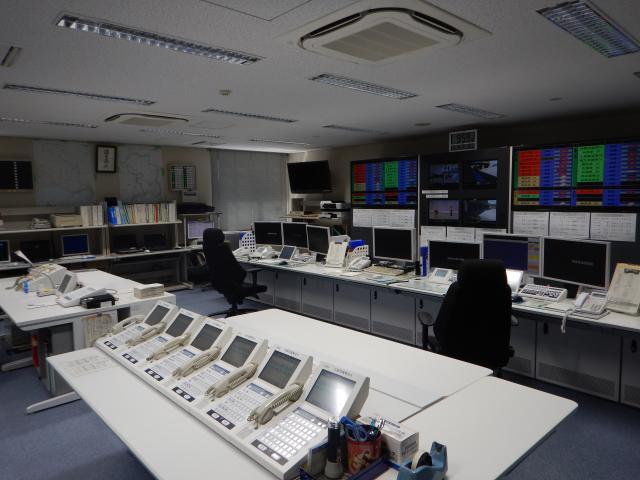 手前に受話器のついた機械、奥にモニターが並んでいる指令室全体を撮影した写真
