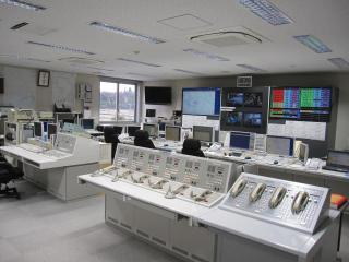 前方に大きなモニターが複数並び、手前に電話機やパソコンなどが並んでいる通信指令室の写真