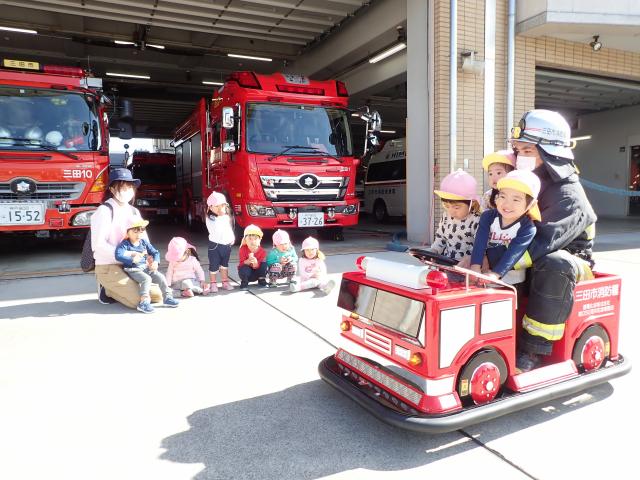 消防署の前に子供たちが並び、消防士と一緒に小さい車に乗っている子どもたちを見ている写真