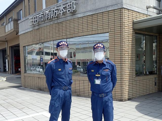 三田市消防本部の建物の前で、二人のフェイスシールドをつけた消防士の男性が並んで立っている写真