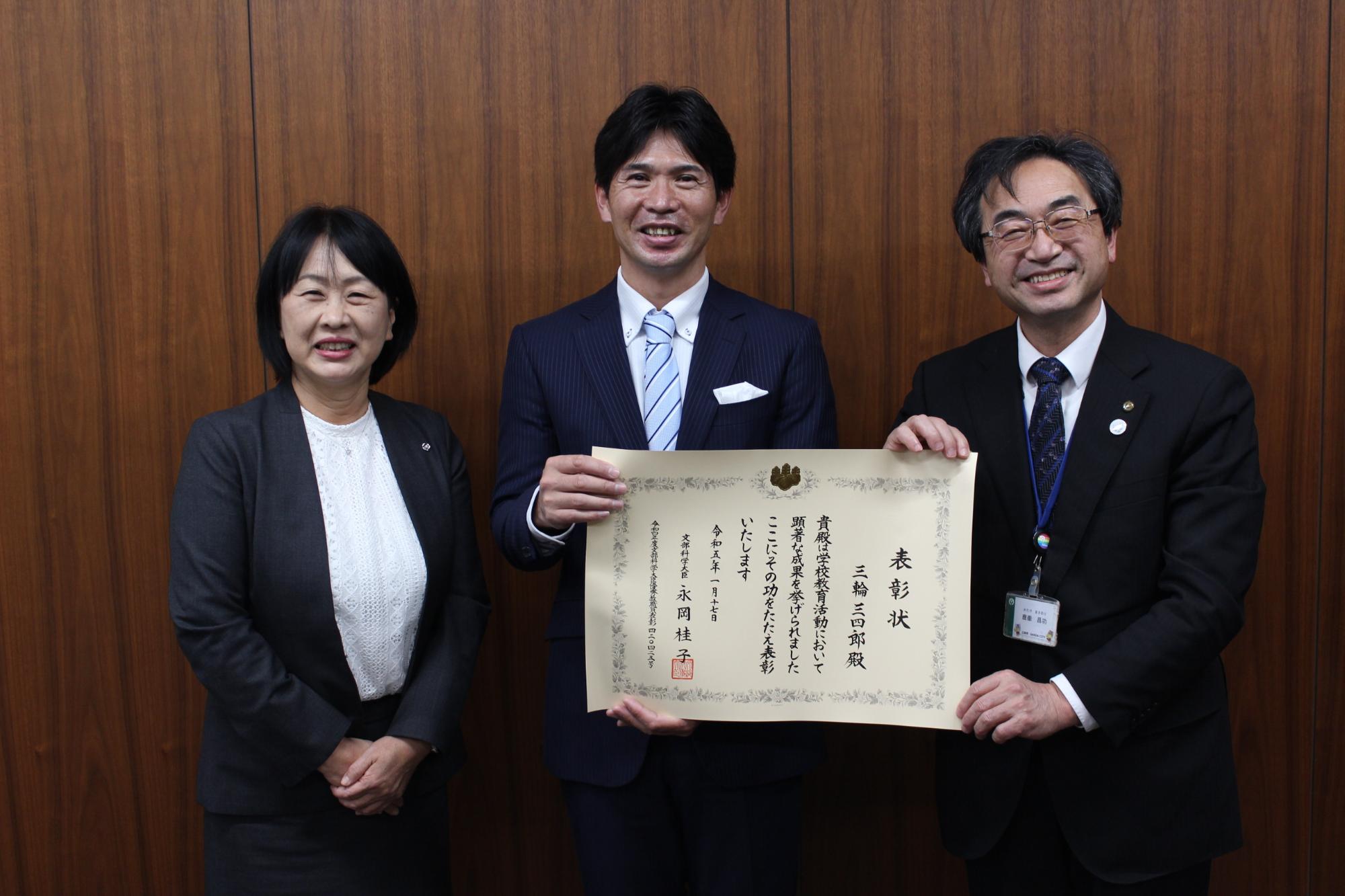 文部科学大臣優秀教職員の表彰状を持つ三輪主幹教諭と、鹿嶽教育長と福本校長が並んでいる写真