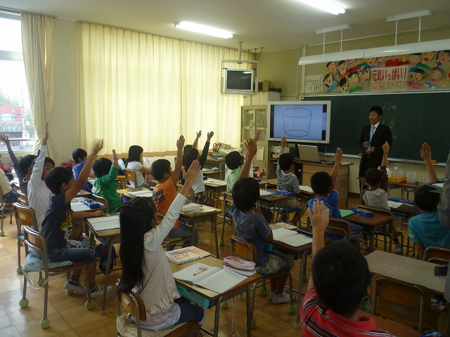 授業で教室前方に立つ教師と手を上げている生徒たちを教室後方から見た写真